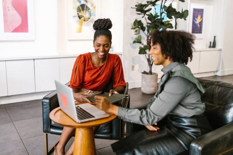 Two professional Black women collaborating in a modern office lounge, smiling and reviewing work on a laptop, with contemporary artwork and indoor plants in the background.