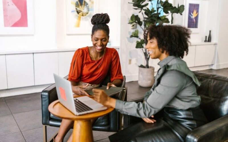 Two professional Black women collaborating in a modern office lounge, smiling and reviewing work on a laptop, with contemporary artwork and indoor plants in the background.